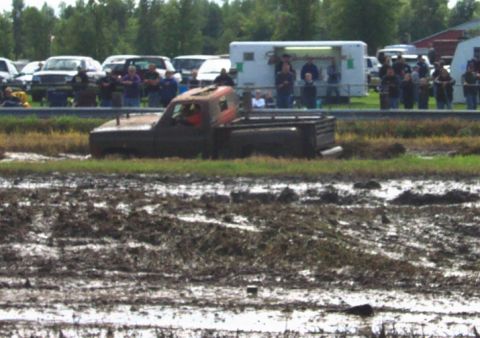 1979 Ford Bronco - A pickup truck is stuck in a mud pit during an event, with spectators watching in the background.