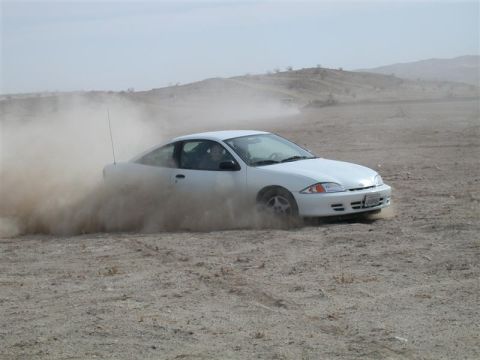 White coupe drifting in dirt, kicking up a large dust cloud. Appears to be a 1995 Chevrolet Cavalier