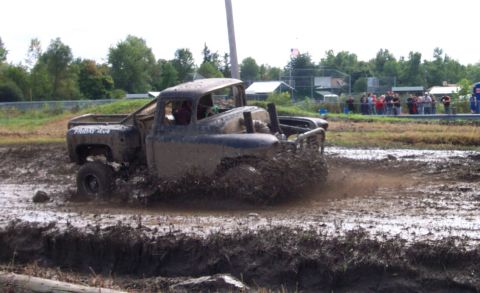 1979 Ford Bronco - A heavily mud-covered pickup truck navigates a challenging mud bog, kicking up a spray of dirt and water.