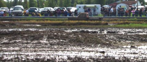1979 Ford Bronco - A truck navigates a muddy track during an event, with onlookers gathered behind a barrier.