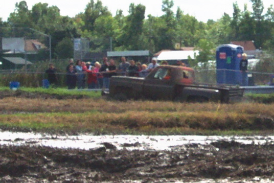 Pickup truck stuck in mud pit with spectators watching. Portable toilet in background