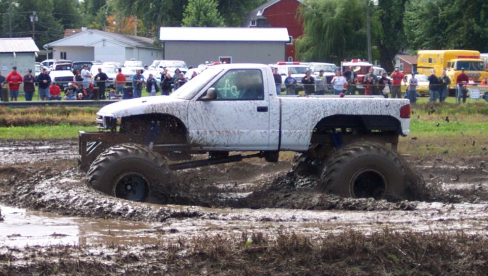 White pickup truck with large tires stuck in a mud pit, spectators in background