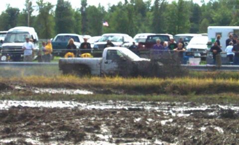 1979 Ford Bronco - A white pickup truck is stuck in a mud pit during an event, with spectators watching from behind a fence.