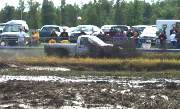 White pickup truck stuck in mud pit with spectators in background