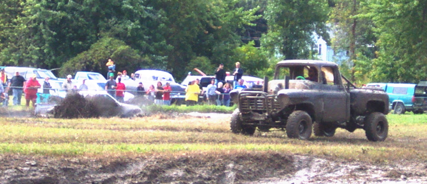 Muddy truck in a field with spectators watching a vehicle stuck in the mud
