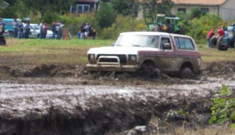 1979 Ford Bronco - A two-tone Ford Bronco is stuck deep in a mud pit during an event, with spectators and other vehicles visible.