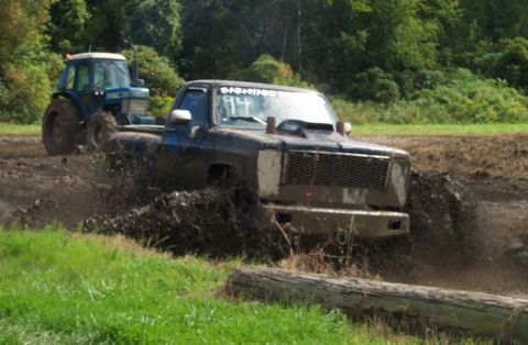 1979 Ford Bronco - A black pickup truck navigates a muddy track, with a blue tractor visible behind it.