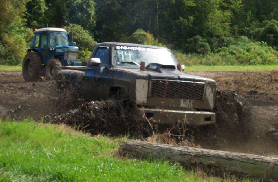Mud-splattered black pickup truck with a hood scoop driving through mud, blue tractor in background