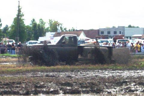 1979 Ford Bronco - A dark pickup truck navigates a muddy obstacle course with spectators in the background.
