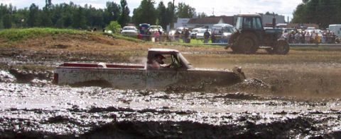 1979 Ford Bronco - Pickup truck navigates a muddy track during an event, with a tractor and crowd visible.