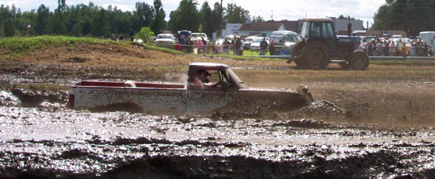 Pickup truck driving through a mud bog with a tractor and spectators in the background