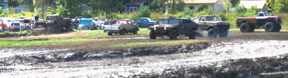 Muddy field with several trucks and an HMMWV driving through mud, spectators in background