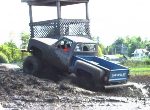 1979 Ford Bronco - A Chevrolet pickup truck navigates a muddy terrain, with a raised wooden platform in the background.