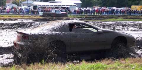1979 Ford Bronco - A sports car navigates a challenging mud pit during an event, with spectators watching in the background.