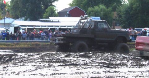1979 Ford Bronco - A heavily modified truck navigates a muddy track, with spectators gathered behind a barrier.