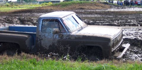 1979 Ford Bronco - A muddy pickup truck is stuck in a mud pit, with spectators in the background. The grille indicates a 1980s model.