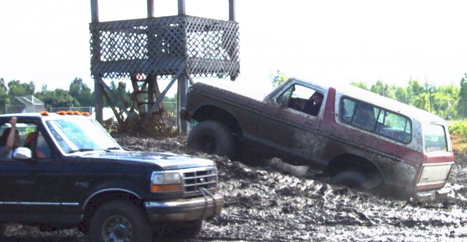 Ford Bronco stuck in mud next to a pickup truck, with a wooden structure in the background