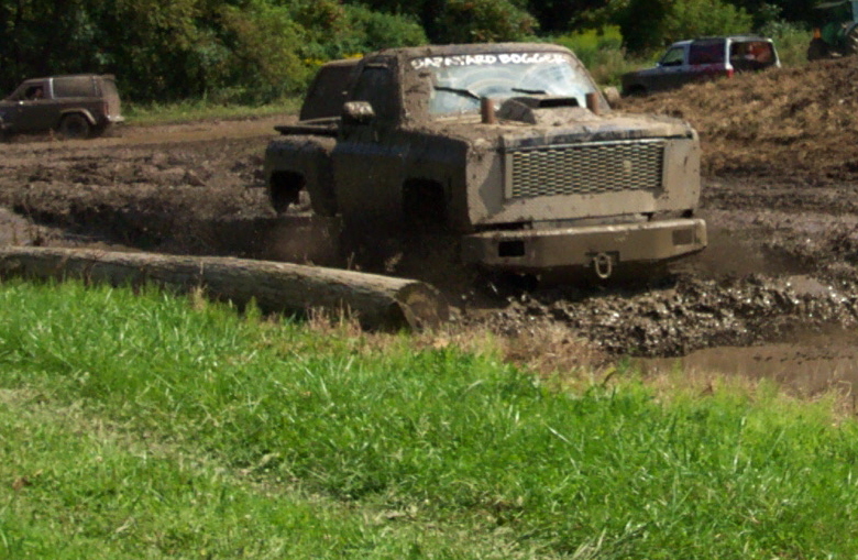 Mud-splattered pickup truck with a large grille and hood scoop stuck in mud, with a log in the foreground