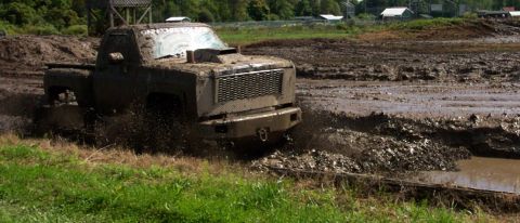 1979 Ford Bronco - A heavily muddied pickup truck navigates a challenging mud bog, kicking up spray as it goes.