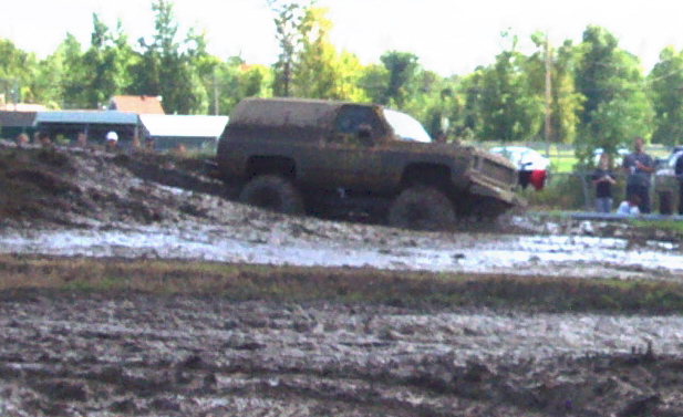 Muddy SUV stuck in a mud pit with trees and people in the background