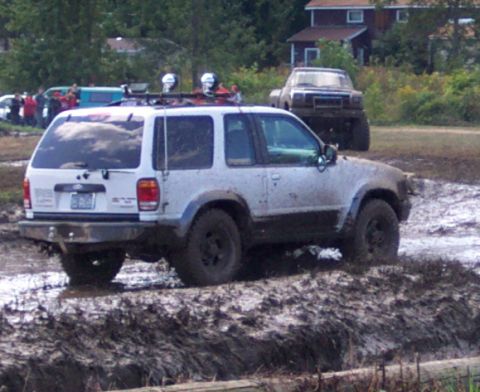 1979 Ford Bronco - White SUV navigates a muddy obstacle course with another vehicle visible behind it.