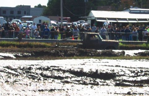 1979 Ford Bronco - A truck navigates a muddy track during an event, observed by a crowd of onlookers.
