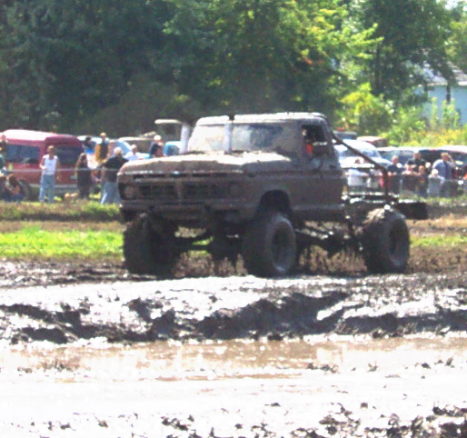 Mud-covered pickup truck with large tires driving through mud, front grille visible