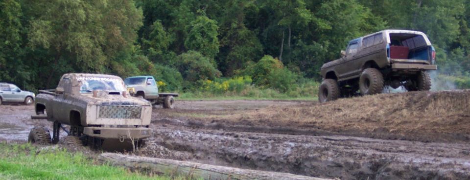 Muddy trucks and a lifted SUV driving through a muddy field with trees in the background
