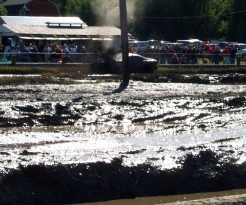 1979 Ford Bronco - A vehicle participates in a mud bogging event, kicking up mud and smoke as spectators watch from a covered area.