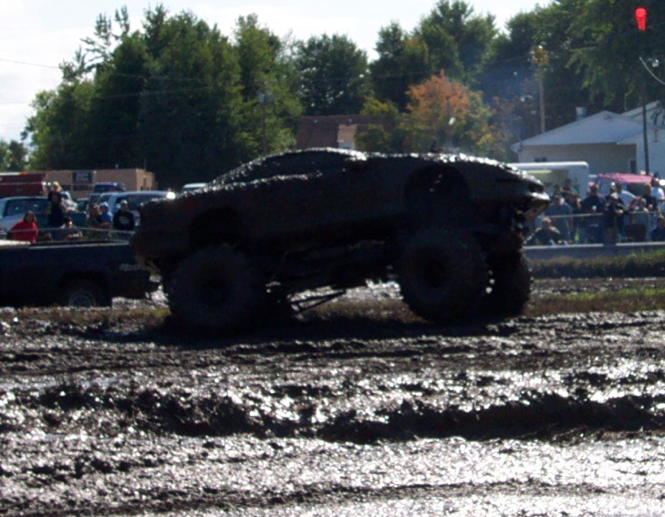 Mud-covered off-road vehicle with large tires driving through a muddy track, spectators in background