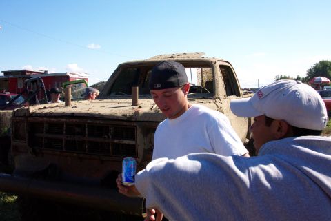 1979 Ford Bronco - Two men stand near a heavily mud-caked truck, with other vehicles and buildings in the background under a clear sky.