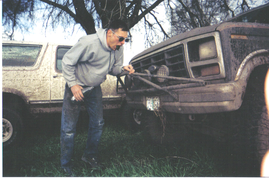 Man in gray hoodie hooking a strap to the front of a mud-caked Ford pickup outdoors