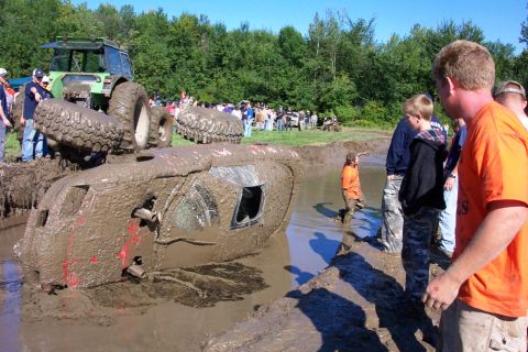 1979 Ford Bronco - Overturned vehicle stuck in mud pit during a bogging event, with a tractor and onlookers present.