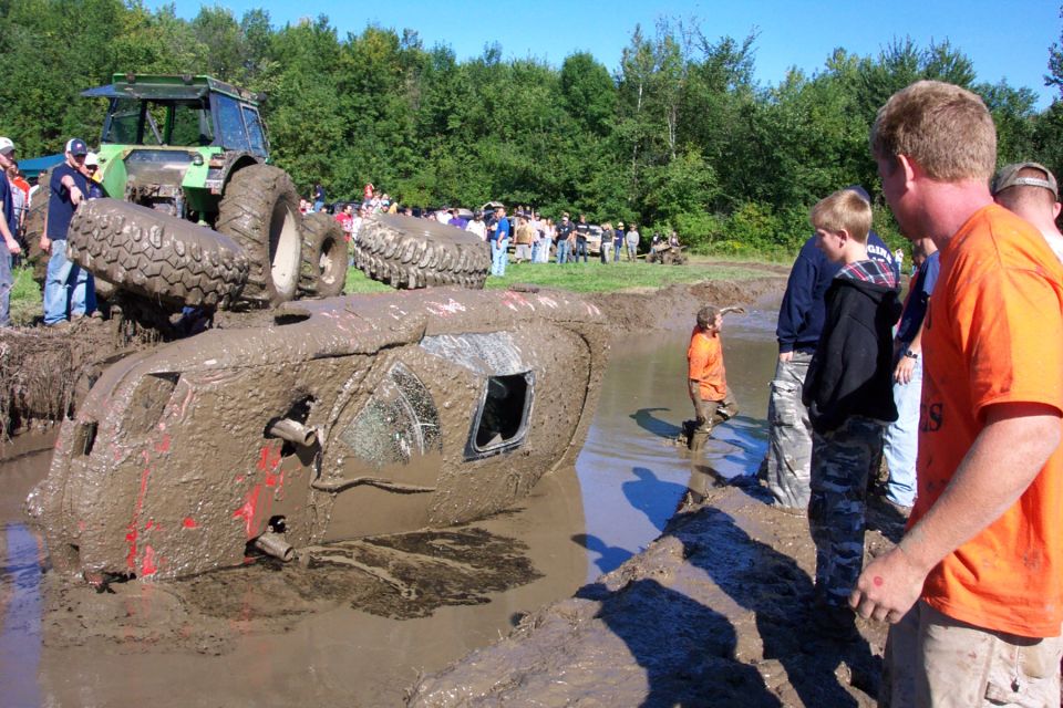 Mud bogging event with an overturned vehicle stuck in the mud, a tractor nearby, and spectators watching