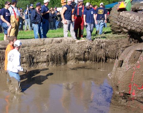1979 Ford Bronco - A heavily mud-caked vehicle is partially submerged in a muddy pit, with onlookers gathered around.