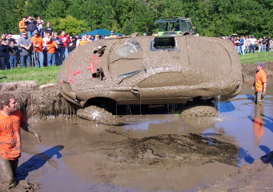 Mud-covered SUV stuck in a bog pit with spectators watching. Another vehicle is visible in the background