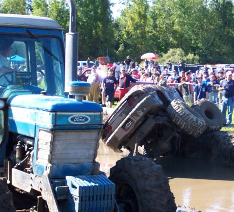 1979 Ford Bronco - An overturned SUV is stuck in a mud pit, with a blue tractor nearby and a crowd watching.