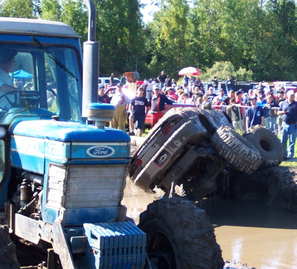 Muddy overturned SUV with Chevrolet badging, stuck in mud pit next to a blue tractor