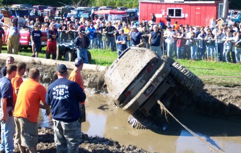 1979 Ford Bronco - An SUV is stuck at a precarious angle in a mud pit during an event, with a crowd of onlookers gathered behind a fence.