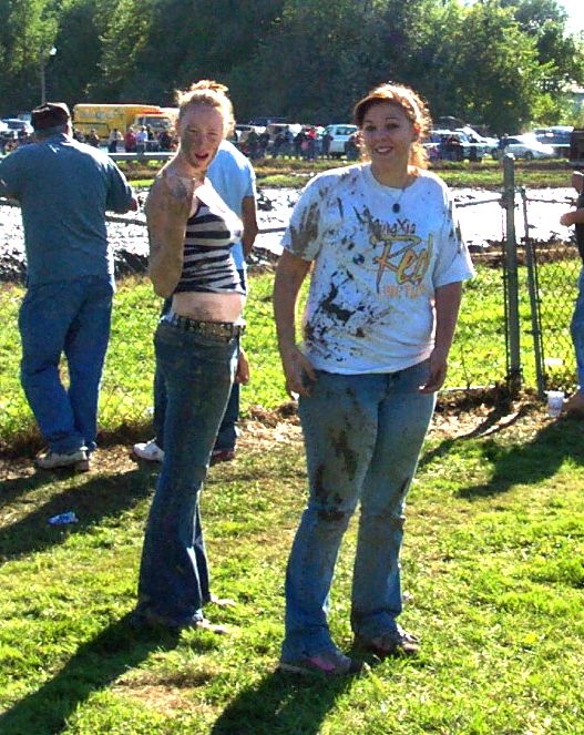 Two women covered in mud stand in a grassy field with vehicles and trees in the background