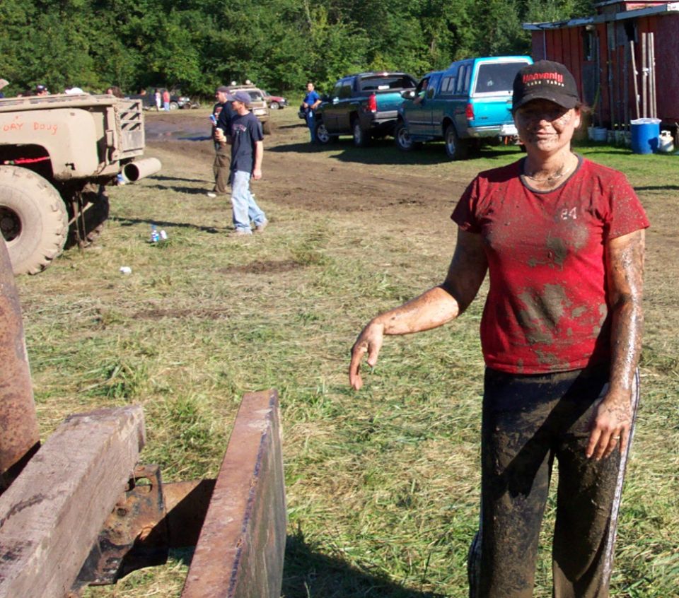 Muddy woman in red shirt and black pants, standing in a field with several vehicles in the background