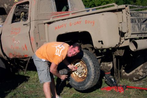 1979 Ford Bronco - A man uses a power tool to work on the wheel of a heavily mud-covered pickup truck, with names scrawled on its doors.