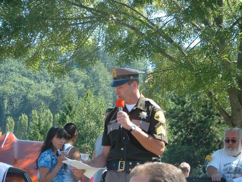 2000 Ford Crown Victoria - A sheriff addresses a crowd at an outdoor gathering, with trees and foliage in the background.