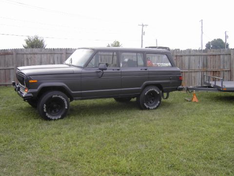 1977 Jeep Wagoneer - A dark-colored Jeep Grand Wagoneer is parked on a grassy lawn next to a wooden fence, attached to a small trailer.