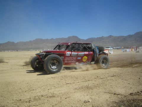 1990 Ford Bronco - dark red off-road race buggy marked number 132 slides through dirt, throwing up a plume of dust on a desert course.