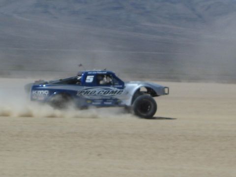 1990 Ford Bronco - blue and silver off-road trophy truck with Pro Comp livery and number 5 speeds across a dusty desert flat.
