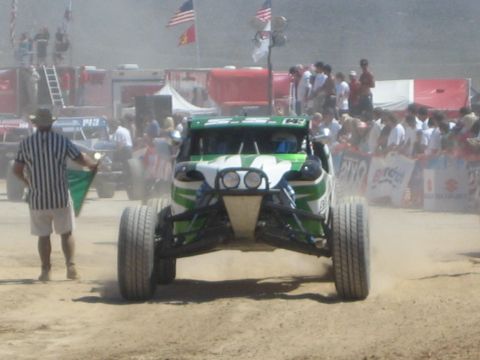 1990 Ford Bronco - green and white off-road race buggy passes a flag-waving marshal as a crowd lines the dusty desert course.