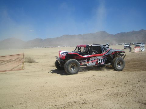 1990 Ford Bronco - red, white and black off-road race buggy marked number 142 races across a dusty desert flat below mountains.