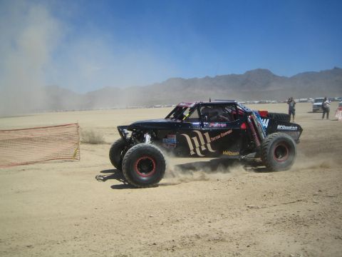 1990 Ford Bronco - black off-road race buggy bearing a Monster Energy logo speeds across a dusty desert flat below distant mountains.