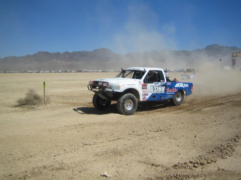 1990 Ford Bronco - white and blue off-road race truck with Voss livery slides through dirt on a dusty desert race course.